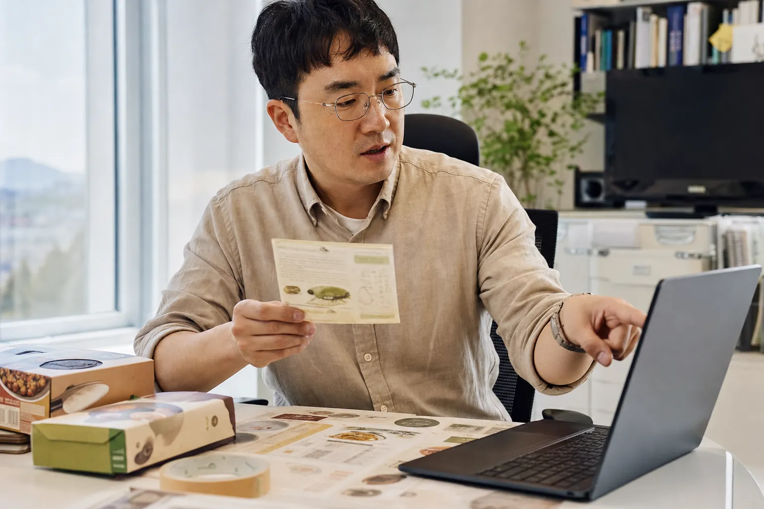 40대 초반 한국인 남성 식품 브랜드 담당자가 제품 포장 라벨을 들고 노트북 화면과 대조하며 확인하는 모습